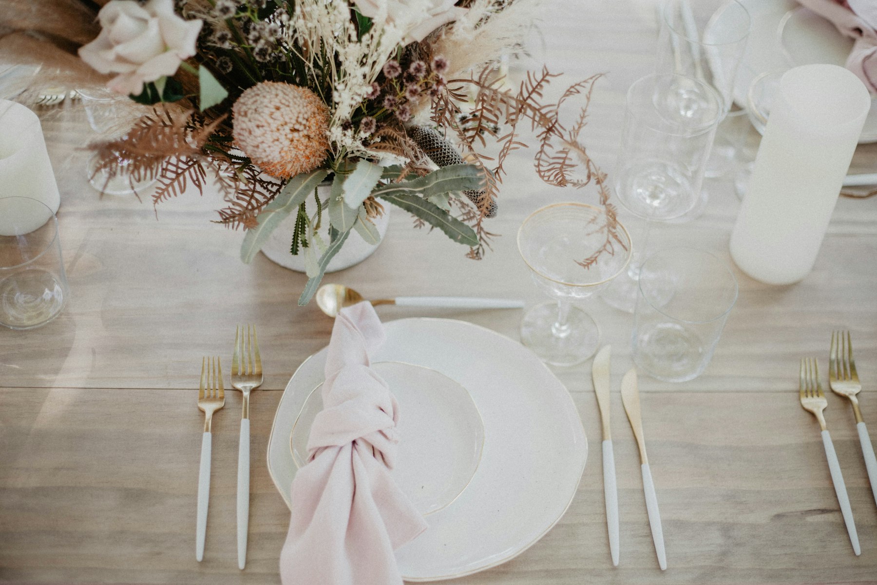 A place setting seen from above: stoneware plate, knotted linen napkin, gold flatware, and dried florals